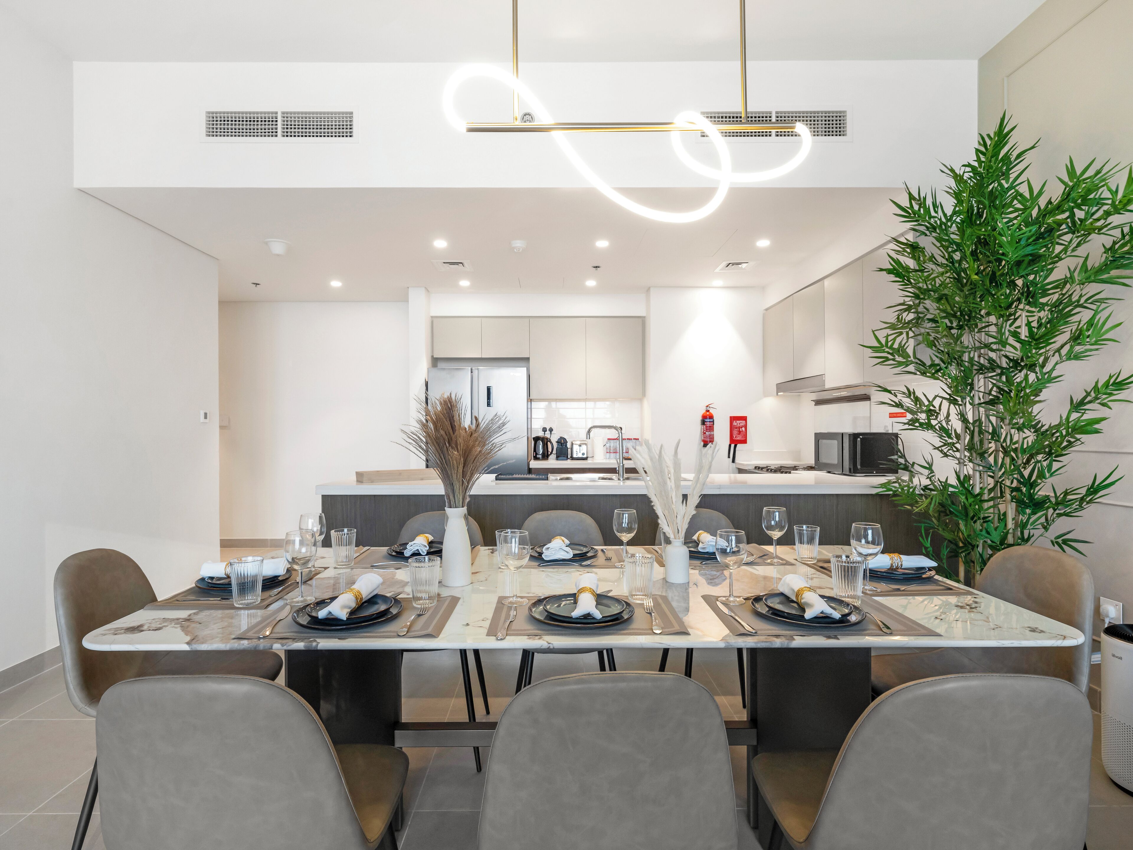 A modern dining area with a long table set for six, a kitchen in the background, and a large green plant on the right.
