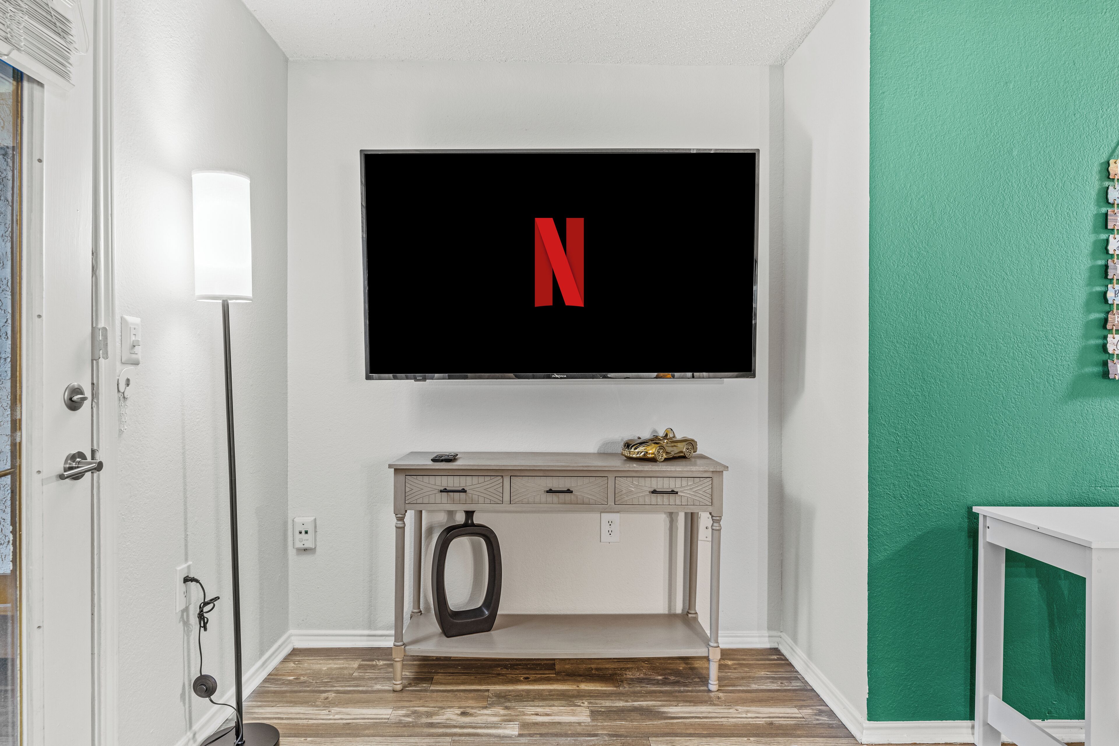 A room with a TV mounted on the wall displaying the Netflix logo, a desk with drawers, a small car model, and a green textured wall.