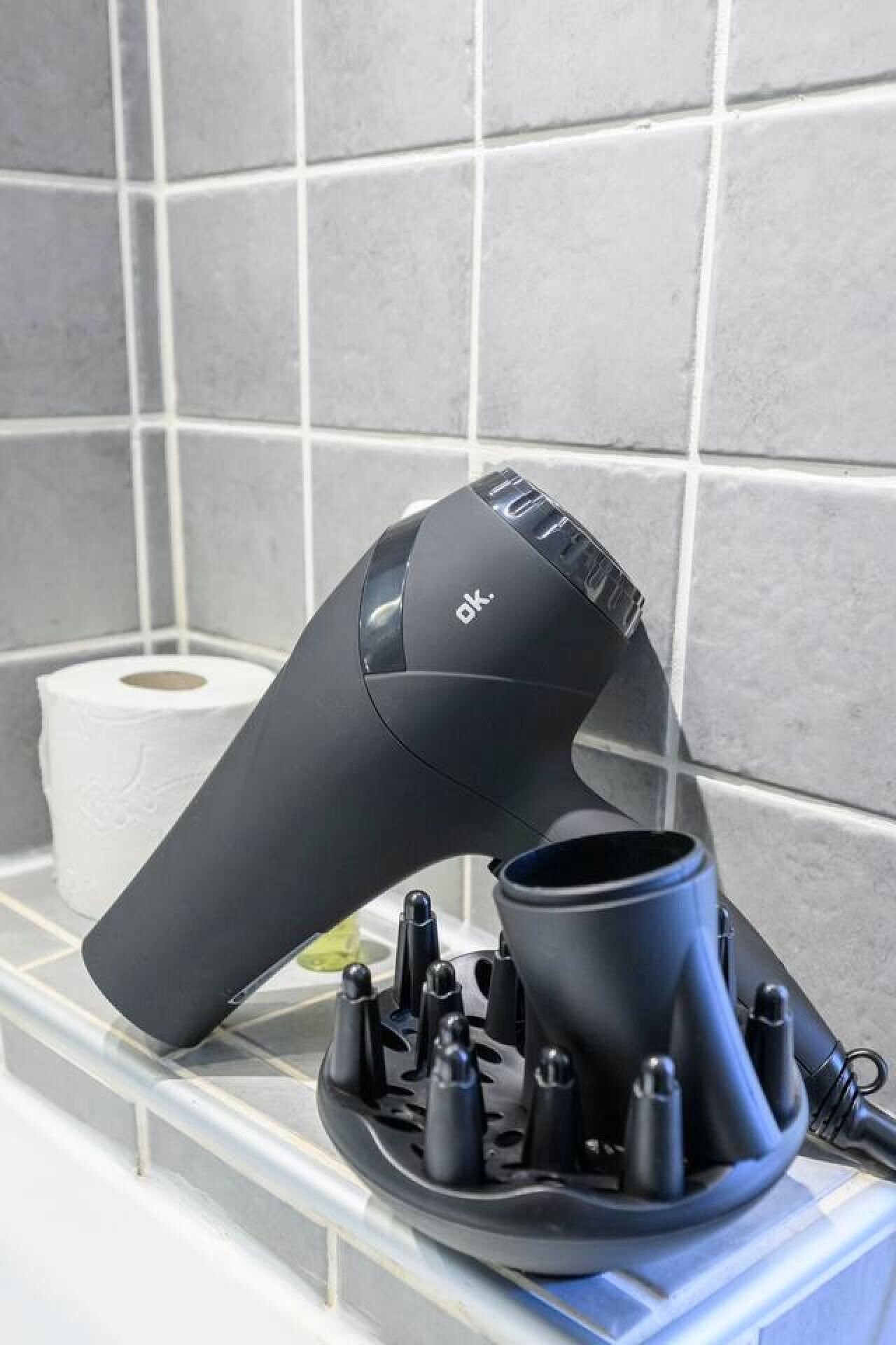 Ein Haartrockner auf einer Badezimmerablage, in der Nähe eine Toilettenpapierrolle.
