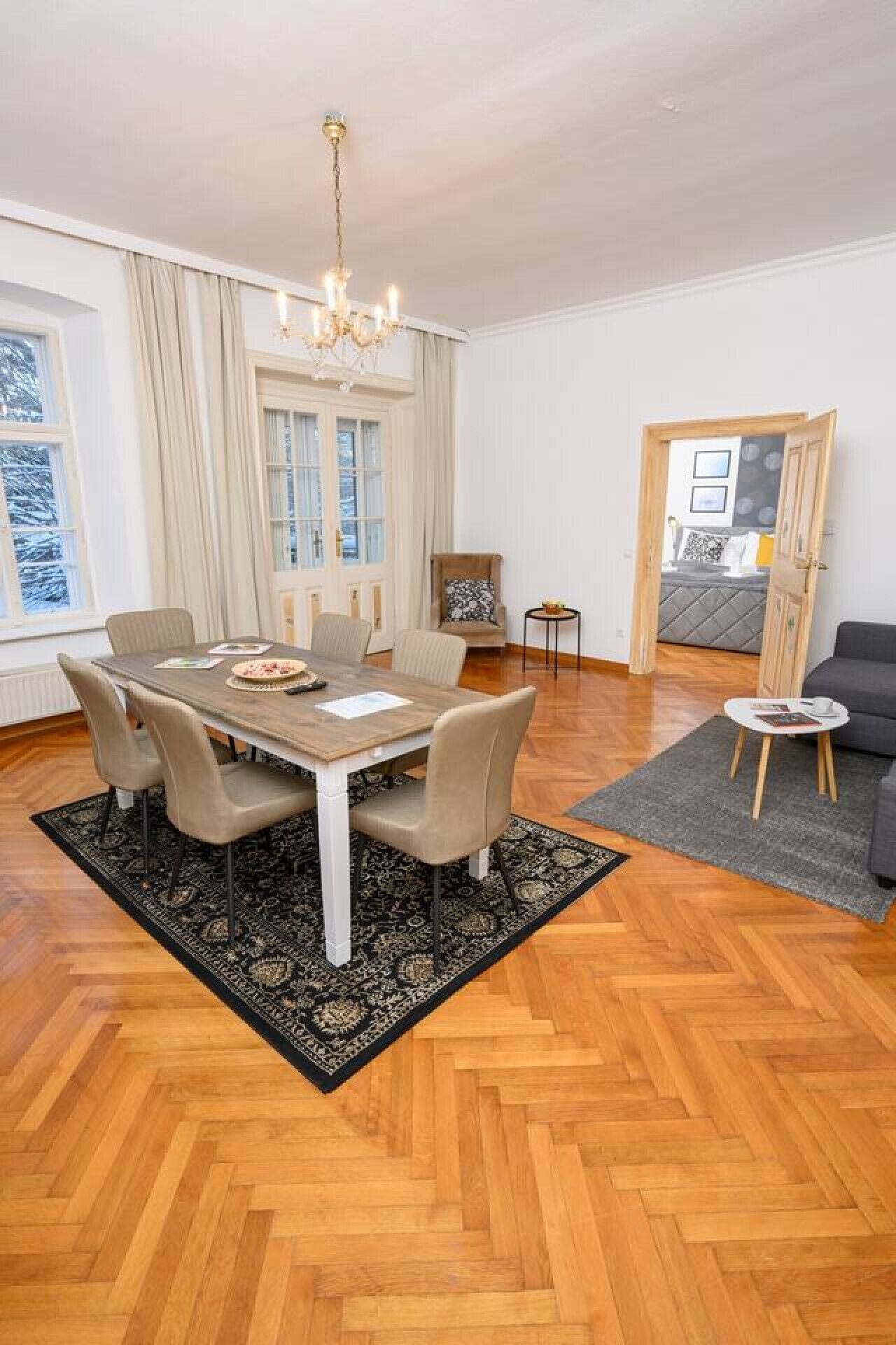 A dining area with a wooden table and chairs, a chandelier, and a view of a snowy landscape through large windows.