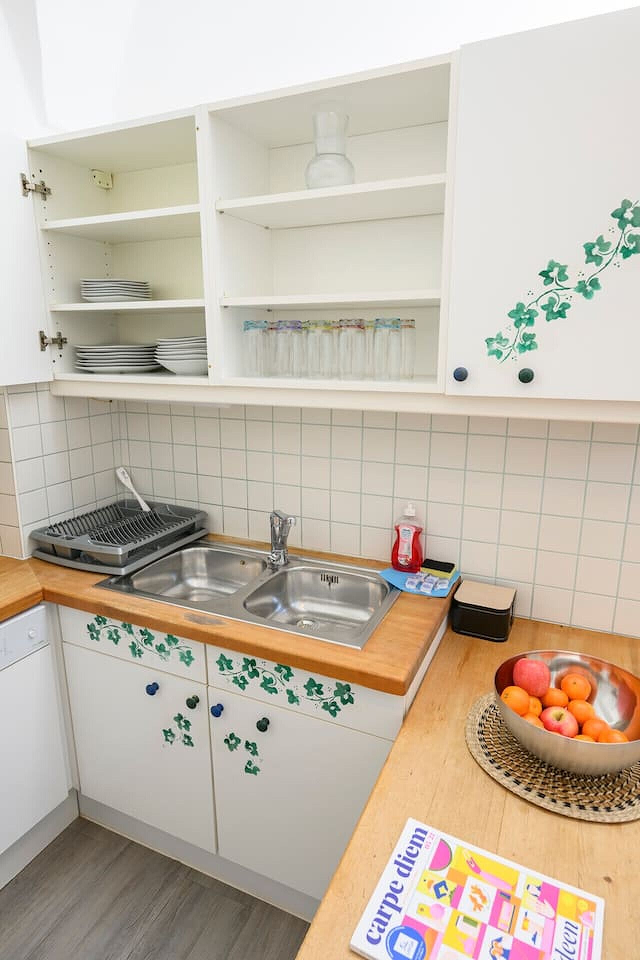 A kitchen with white cabinets, a stainless steel sink, and a wooden countertop. There is a bowl of fruit on the counter and a magazine titled "Carpe Diem".