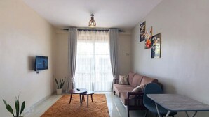 A living room with a sofa, a coffee table, a TV mounted on the wall, and a window with curtains. - Maryz Secret Haven, cozy one bedroom Air BnB Condo (Kampala)
