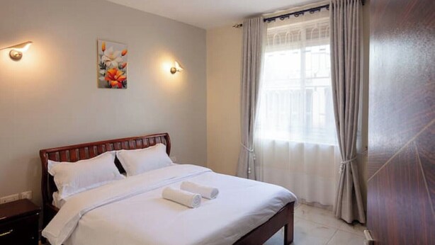 A neatly made bed with white linens and two rolled towels, a wooden headboard, and a painting on the wall. - Maryz Secret Haven, cozy one bedroom Air BnB Condo (Kampala)