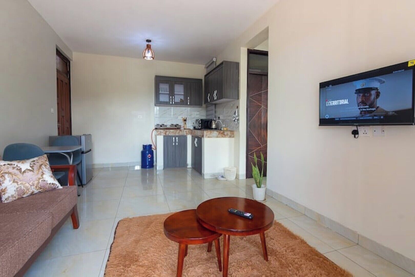 A living room with a sofa, two coffee tables, a TV mounted on the wall, and a kitchen in the background.