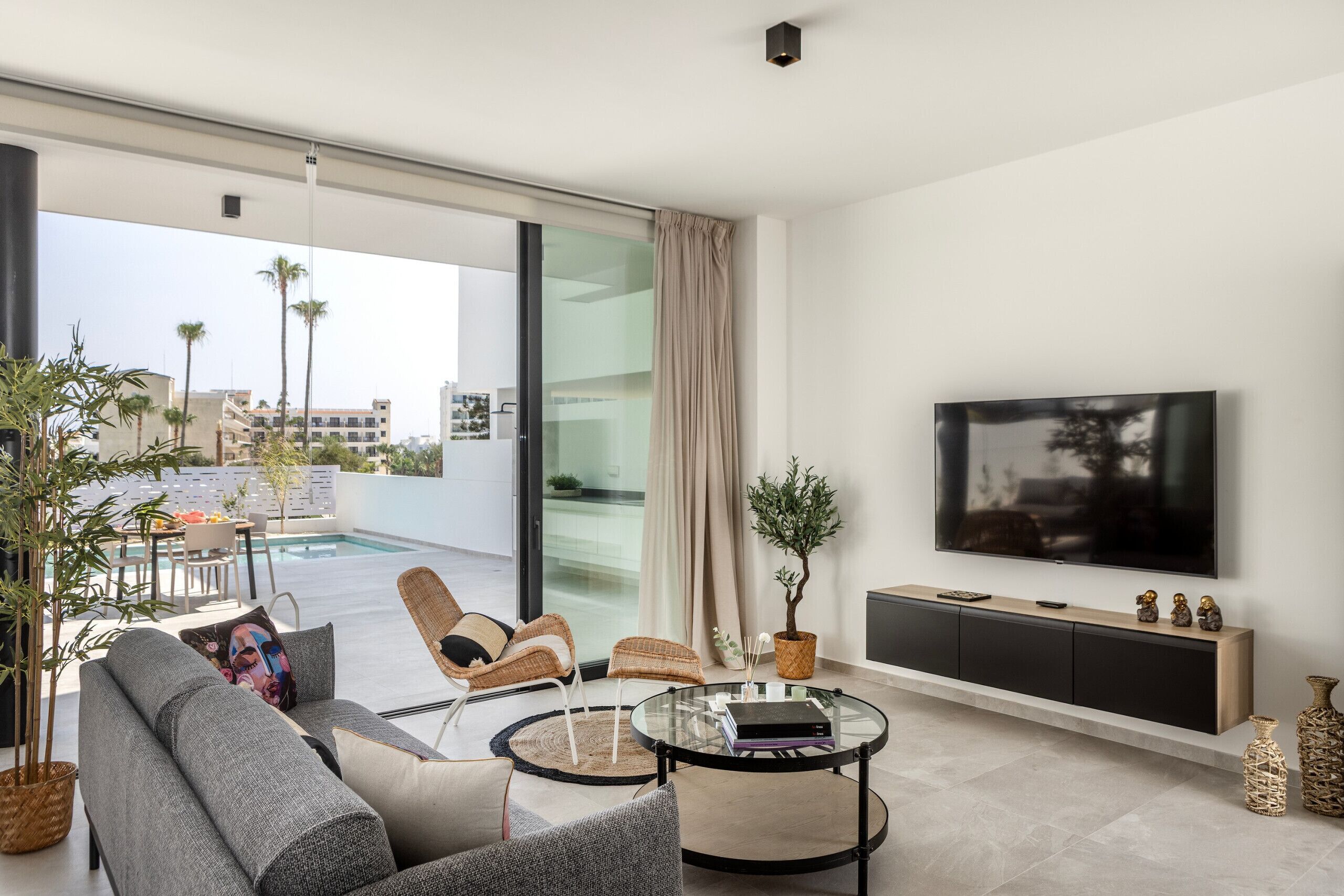 Een moderne woonkamer met een glazen salontafel, grijze banken, rieten stoelen, een flatscreen-tv en uitzicht op een zwembad.