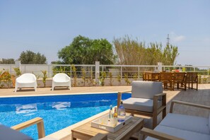 Área da piscina com espreguiçadeiras, uma mesa com bebidas e vista para árvores e uma cerca.