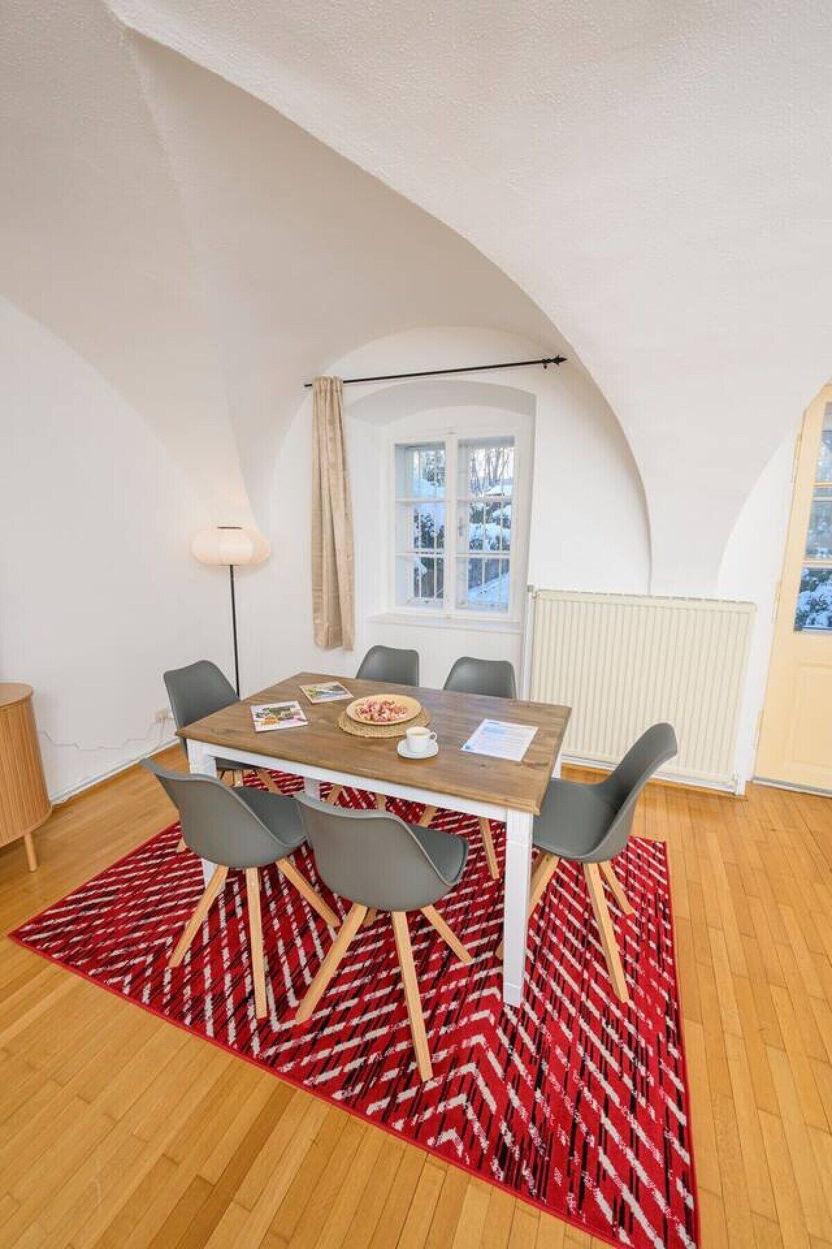 A dining area with a wooden table, grey chairs, and a red patterned rug.