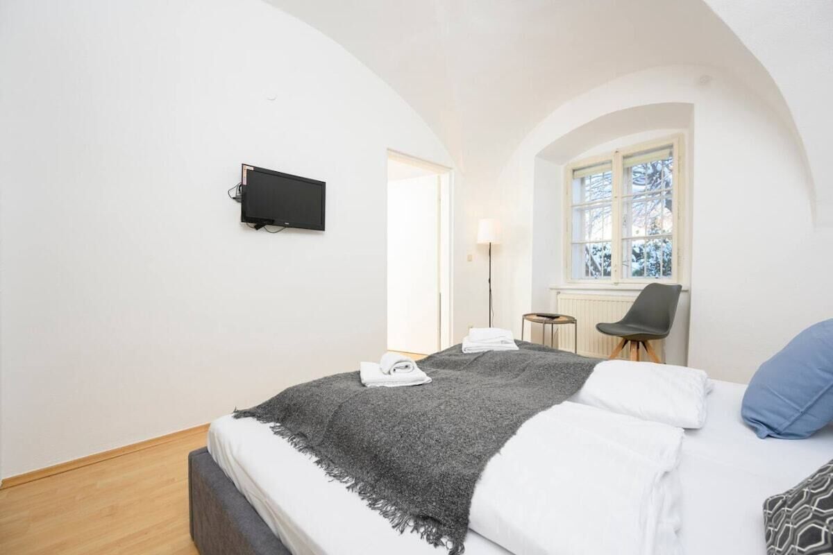 A bedroom with a bed, a TV mounted on the wall, a window with a view, and a chair.