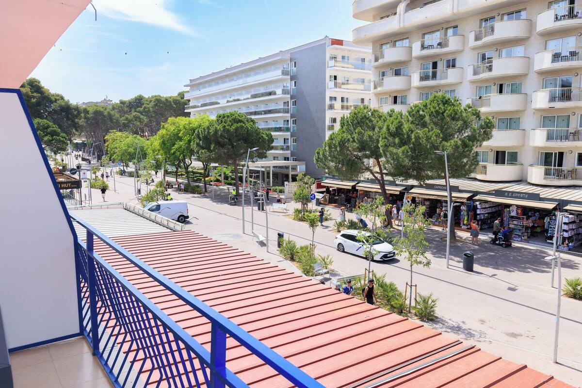 A multi-story hotel with balconies, a covered outdoor seating area, and a street view with shops and parked cars.