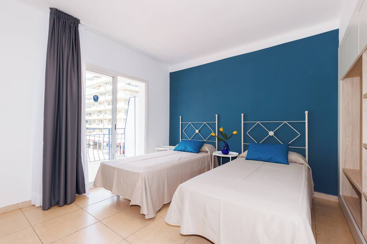 A hotel room with two beds, a blue accent wall, a window with curtains, and a view of a balcony.