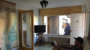 A room with a large map on the wall, a television on a small table, and a person sitting on a couch reading a book. - Phoenix Hostel (San Carlos de Bariloche)