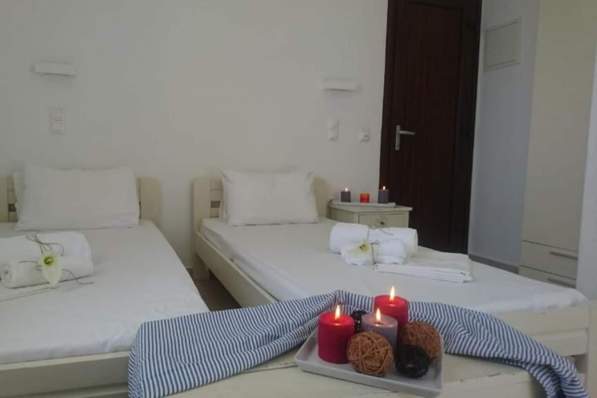 Two single beds with white linens, a small bedside table with a red candle, and a tray with candles and a flower.
