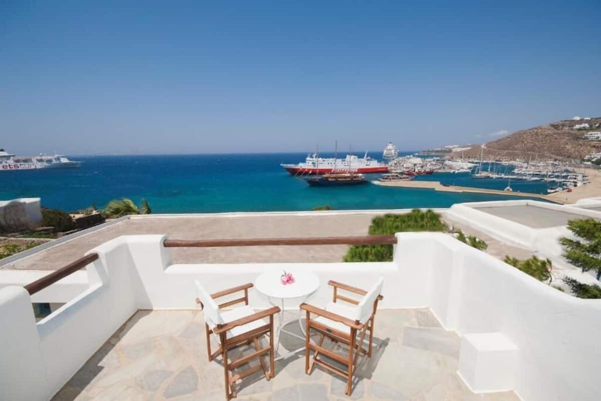 A terrace with two wooden chairs and a small table, overlooking a marina with boats and a beach.