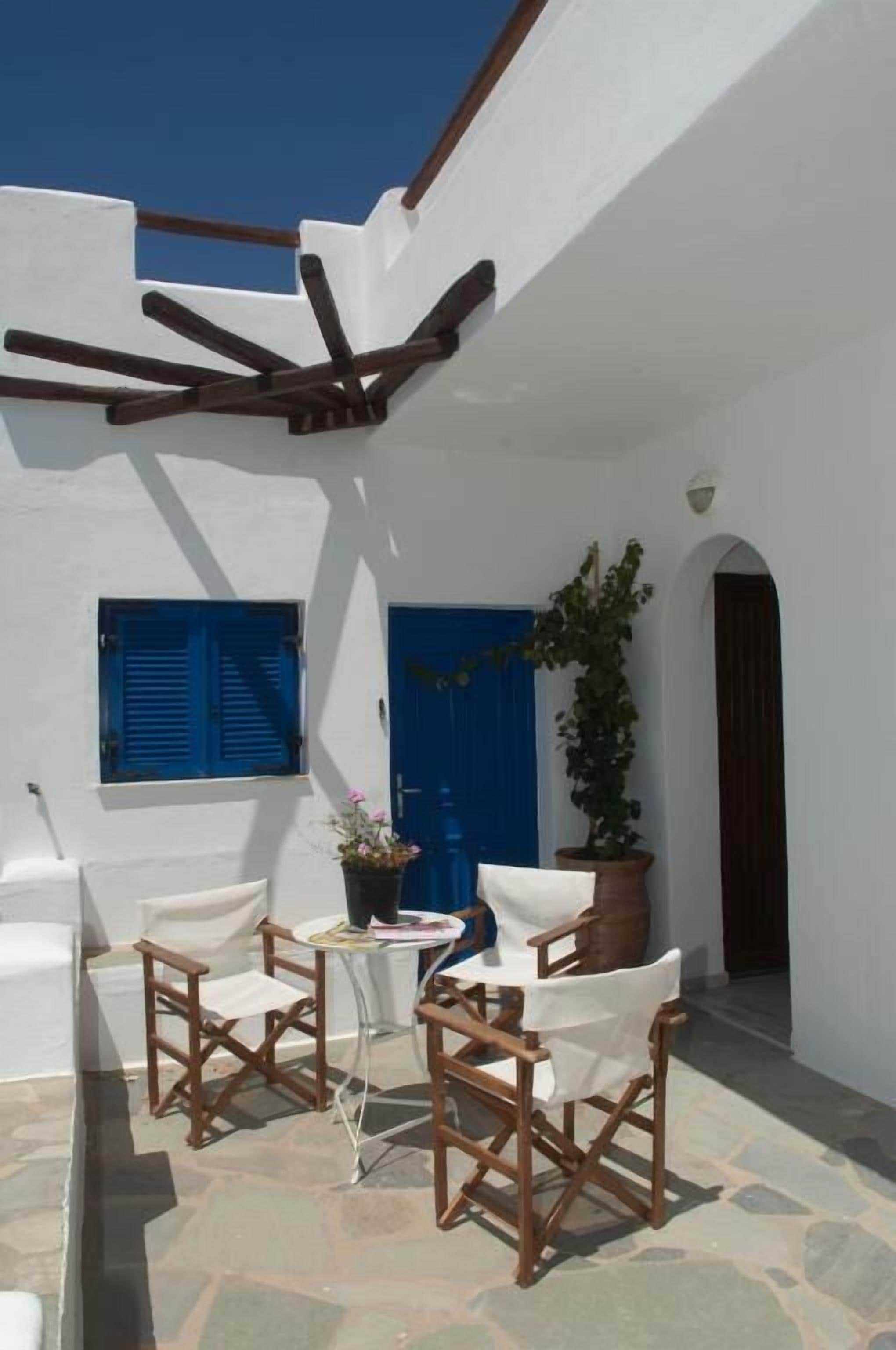 Un espace extérieur aux murs blancs, avec une porte bleue, une plante en pot et des chaises en bois disposées autour d’une petite table.