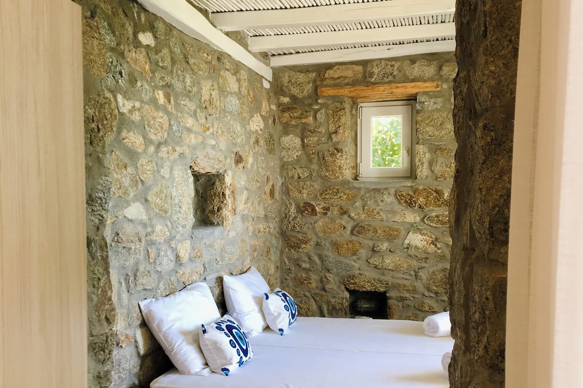 A stone-walled bedroom with a bed, pillows, a small table with candles, and a window.