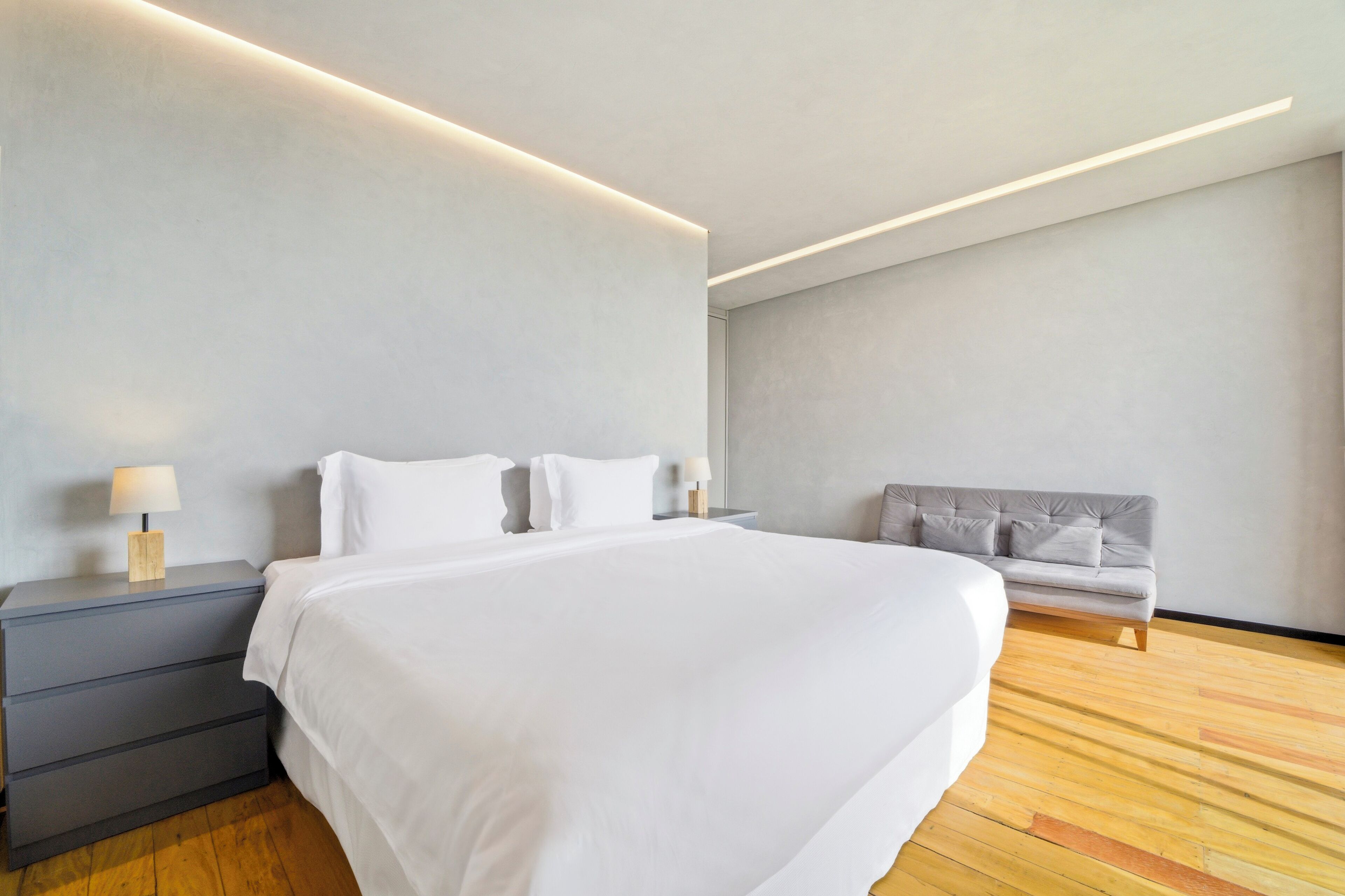 A modern bedroom with a large bed, bedside tables, a sofa, and a wooden floor.