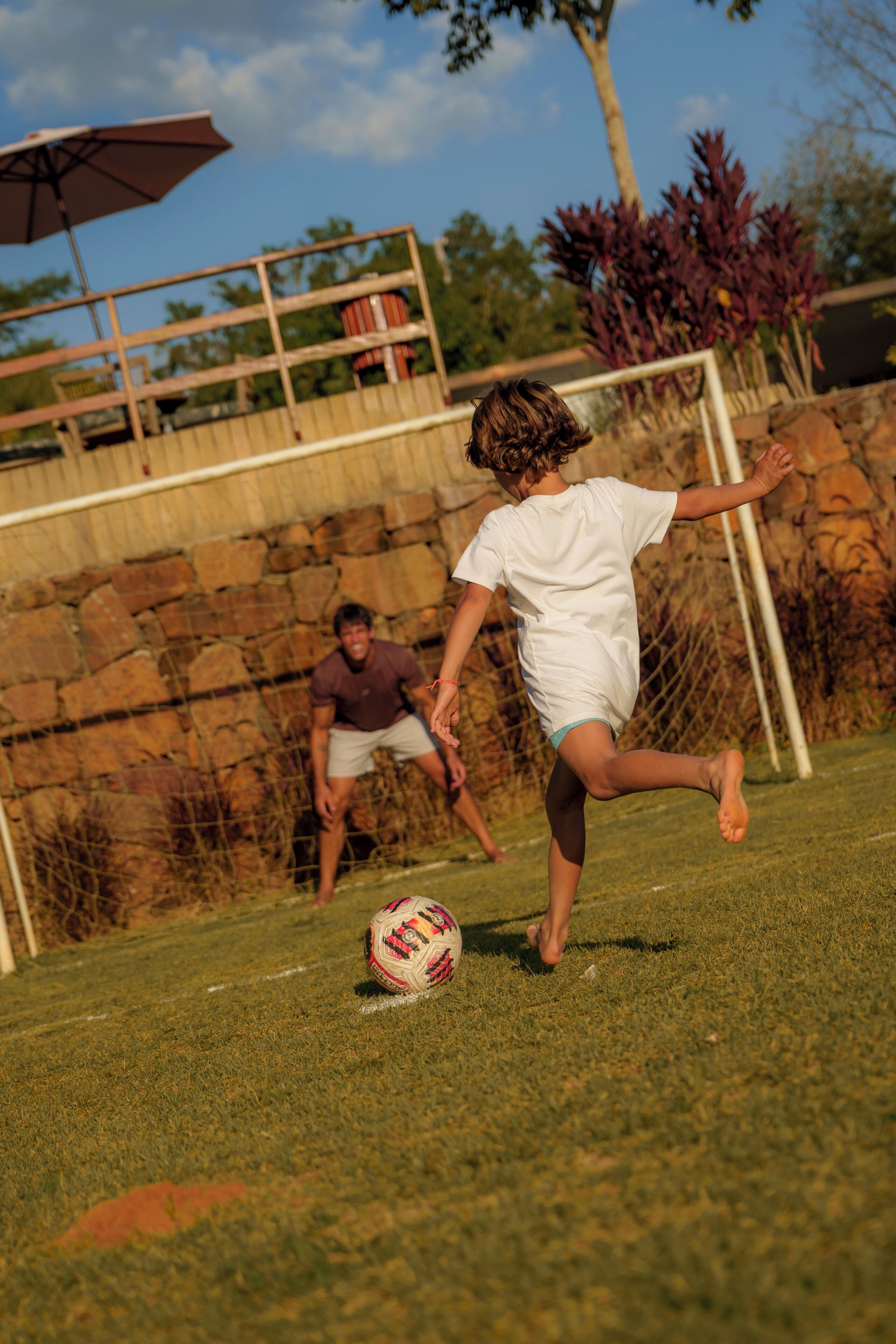 A grassy outdoor area with a goalpost, a soccer ball, and a person kicking the ball.