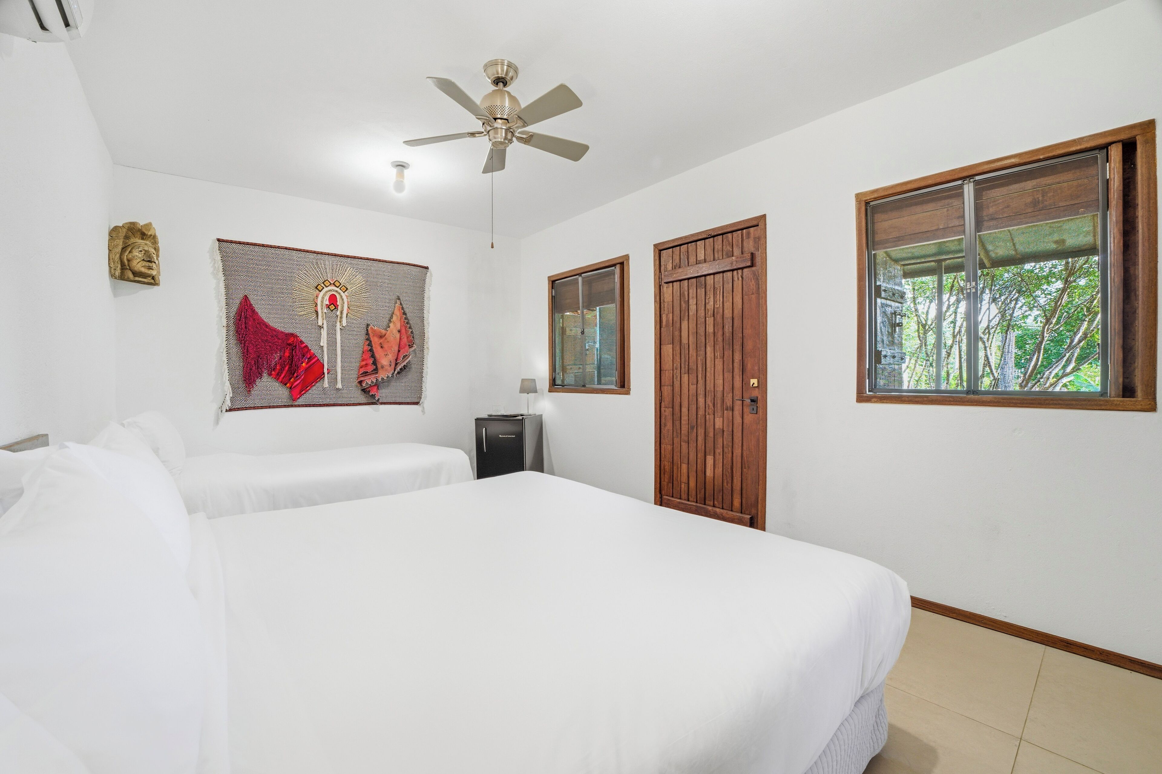 A hotel room with two beds, a ceiling fan, a window with a view, and a wall-mounted artwork.
