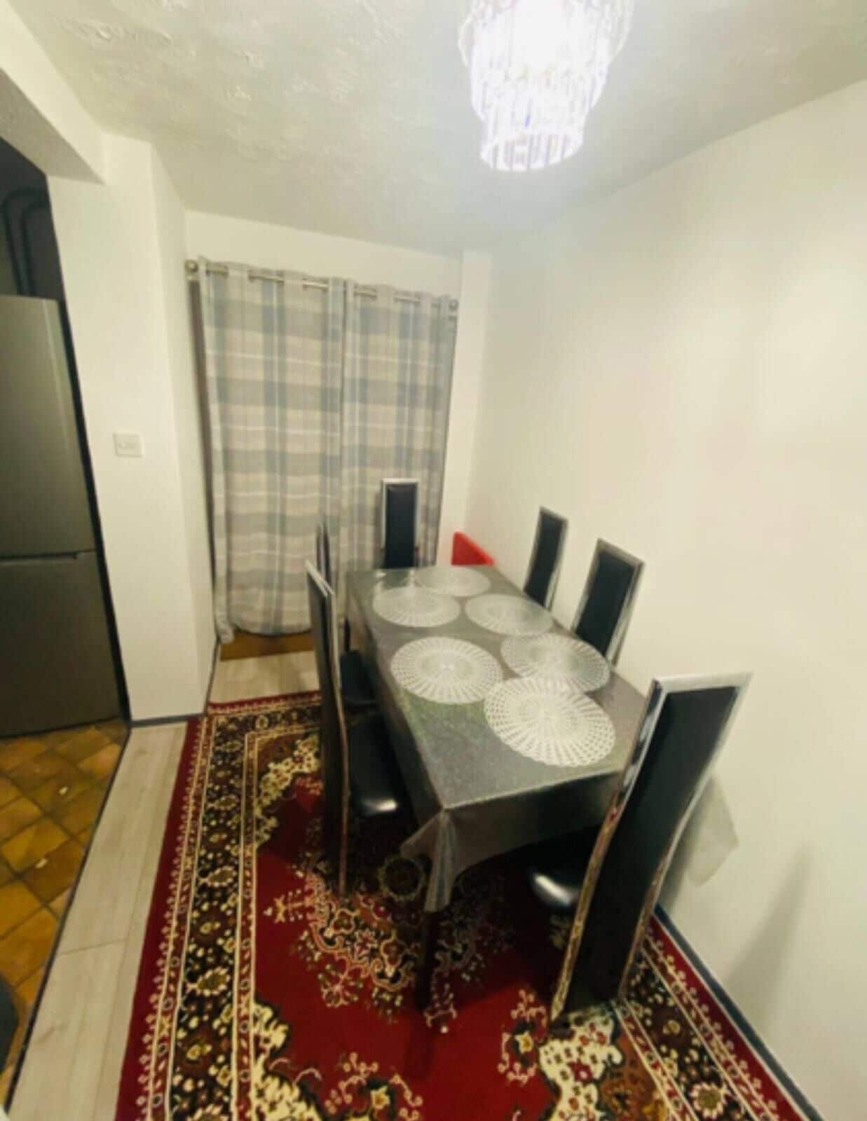 A dining area with a table, chairs, a patterned rug, a refrigerator, and a curtain.
