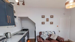 A compact kitchen with a built-in oven, a coffee maker, and a sink. There is a brown leather sofa with a patterned throw blanket and pillows. The room has a wooden floor and white walls. - Harbour View (Whitby)