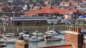 A marina with boats docked, a brick building with a flat roof, and a town with red-roofed houses in the background. - Harbour View (Whitby)