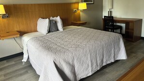 A neatly made bed with a patterned bedspread, two bedside lamps, a wooden headboard, a desk with a chair, and a framed picture on the wall.