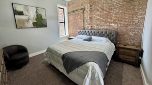 A bedroom with a brick wall, a tufted headboard, a large bed, a chair, a nightstand, and a ceiling fan. - Loft Living - Small Town Charm 
3-bedroom loft in beautiful Downtown Litchfield (Litchfield)