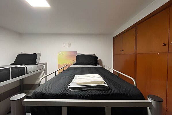 A dormitory room with two single beds, a wooden wardrobe, a poster on the wall, and a small trash bin.