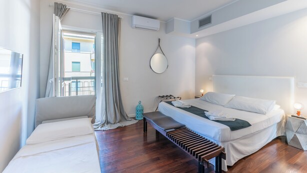 A modern hotel room with a large bed, a bench, a small table, and a window with curtains. - VATICAN PIXEL APARTMENTS®️ (Roma)