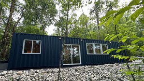 A modern, dark blue, container-style building with large windows, surrounded by a rocky landscape and dense forest.