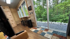 A cozy cabin interior with a wooden interior, a sofa, a dining area, and a large window offering a view of a forest.