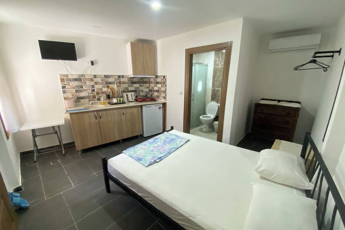 A compact hotel room with a bed, a small kitchenette, a bathroom, and a TV mounted on the wall.
