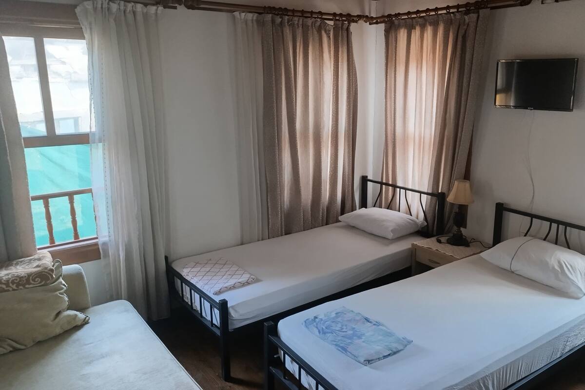 A room with two single beds, a TV mounted on the wall, and a window with curtains.