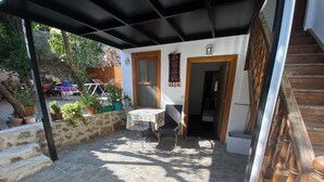 Eine überdachte Terrasse im Freien mit Steinboden, einem Tisch und Stühlen. Es befinden sich Blumentöpfe und eine Holztreppe dort.