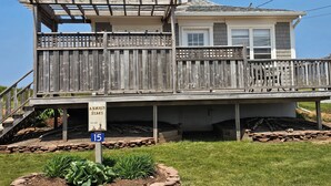 A two-story house with a wooden deck, a covered porch, and a stone foundation. - Cozy Cottage 50 meters from the Ocean (North Rustico)