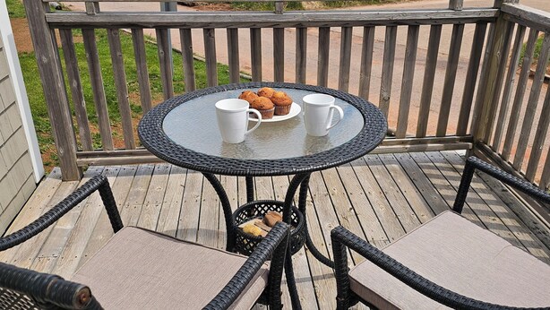 A wooden deck with a round table and two chairs, a basket with items, and a view of the ocean. - Cozy Cottage 50 meters from the Ocean (North Rustico)