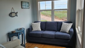 A living room with a dark blue sofa, a white armchair, a ceiling fan, and a large window with a view of a road and greenery. - Cozy Cottage 50 meters from the Ocean (North Rustico)
