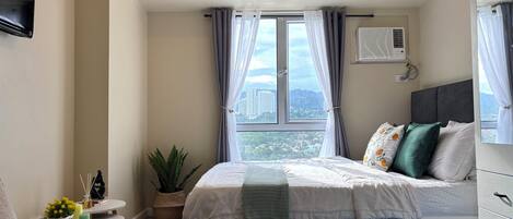 A bedroom with a bed, a bedside table, a potted plant, a window with curtains, and a wall-mounted air conditioner.