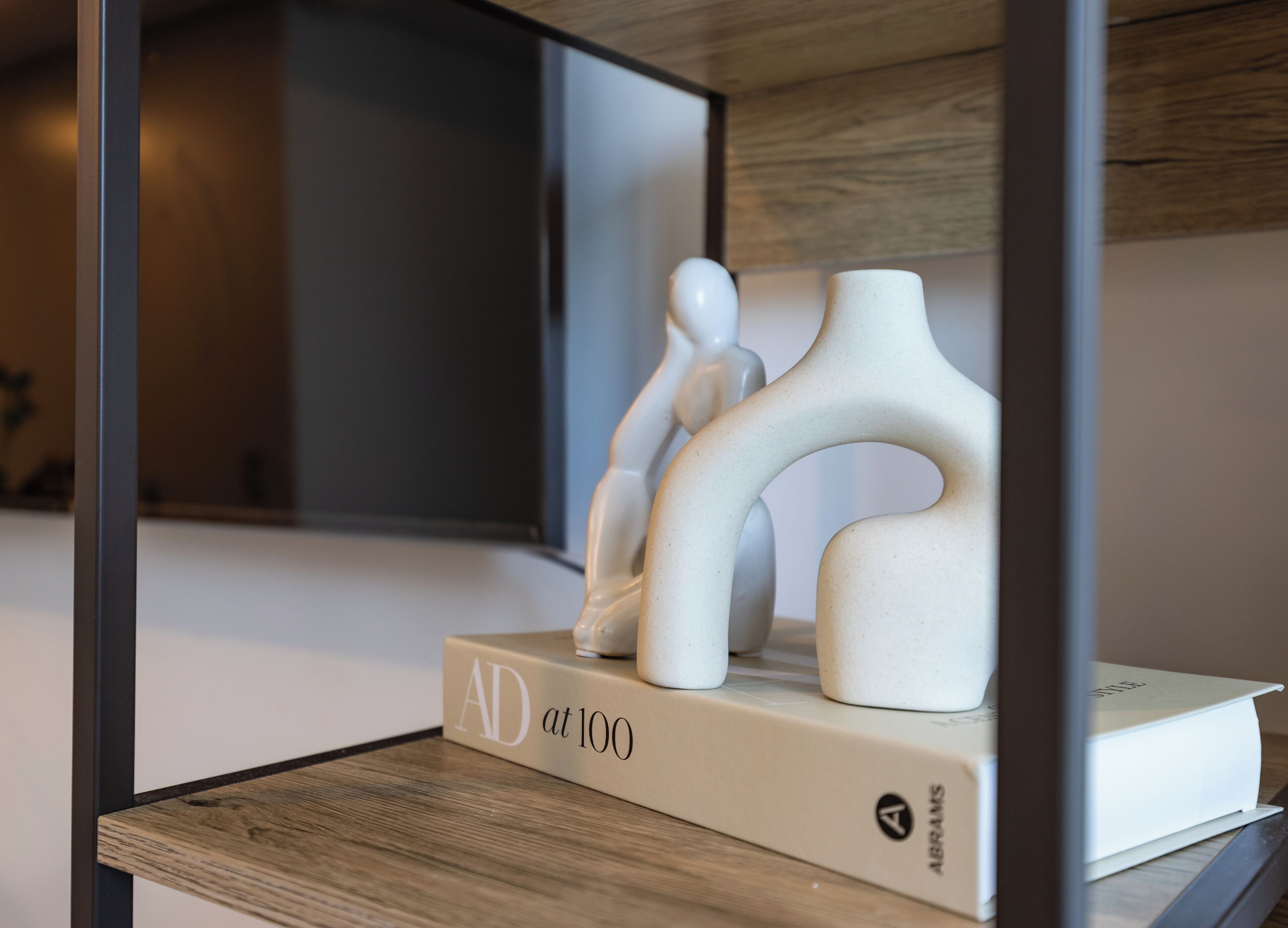 Ein modernes Interieur mit minimalistischem Design, das ein Holzregal mit einem weißen Buch mit dem Titel 'AD at 100' sowie eine weiße Skulptur zeigt.