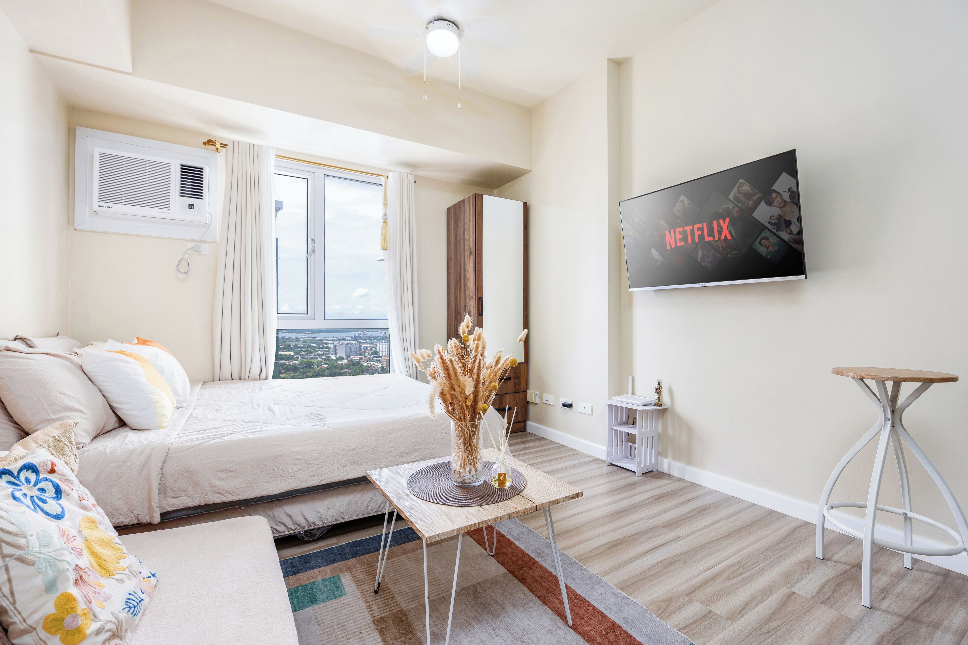 A modern hotel room with a bed, a small table with a vase, a TV on the wall, and a view of the city.