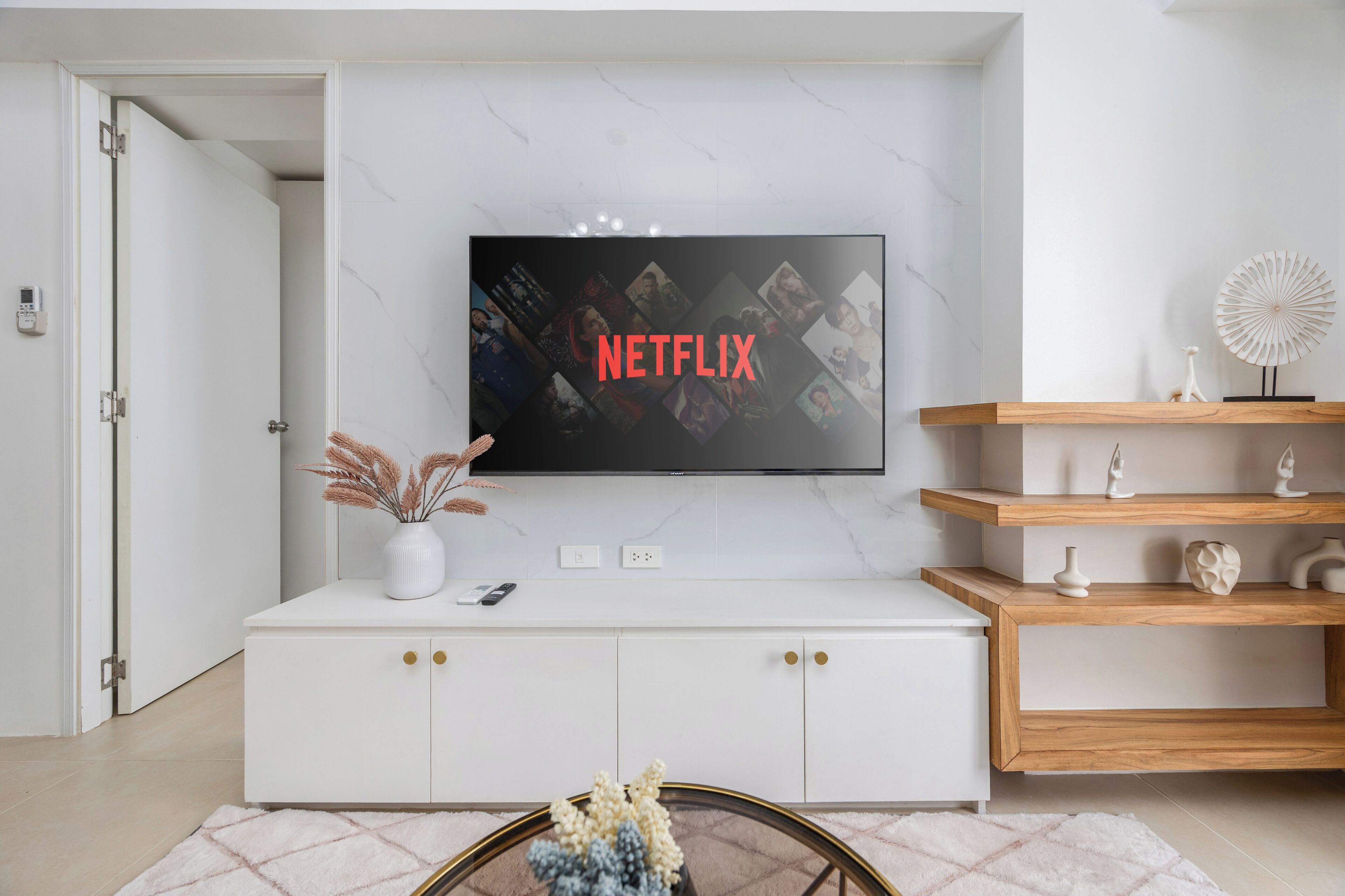 A modern living room with a white cabinet, a mounted TV displaying the Netflix logo, and a wooden shelf with decorative items.