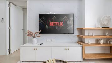 A modern living room with a white cabinet, a mounted TV displaying the Netflix logo, and a wooden shelf with decorative items.
