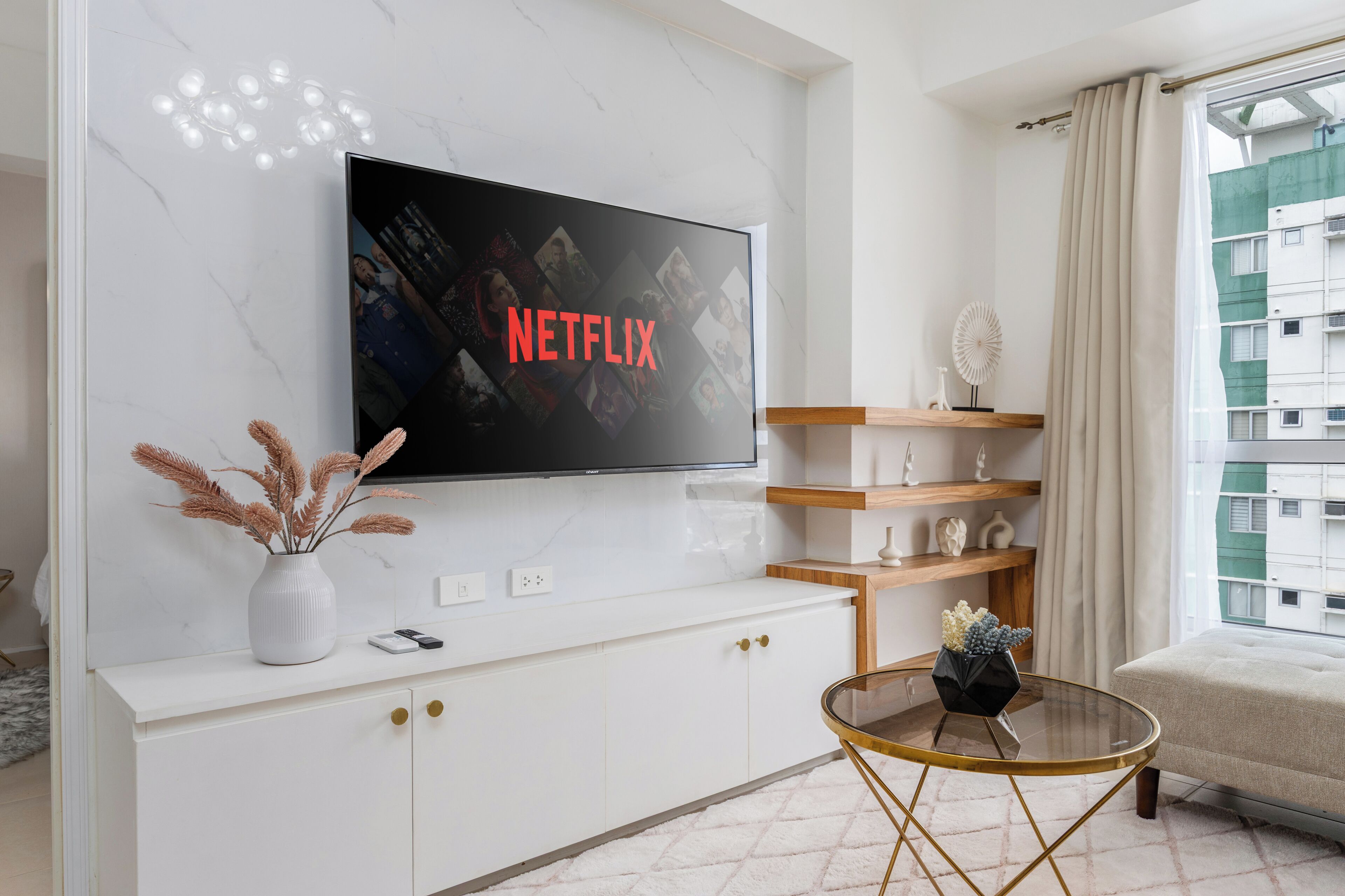 Un salon moderne avec un téléviseur à écran plat fixé à un mur blanc, une table basse en verre, une console blanche avec des poignées dorées et une vue sur un immeuble par la fenêtre.
