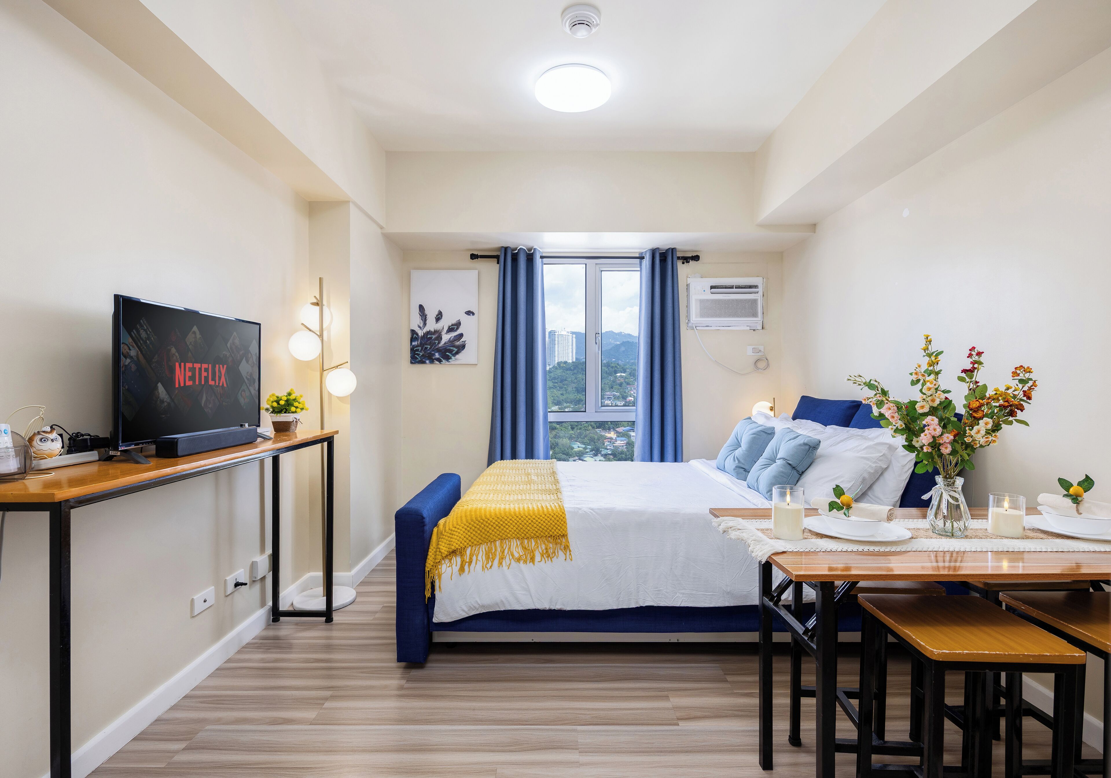 A hotel room with a bed, bedside table, TV, and a dining area with a table and chairs.