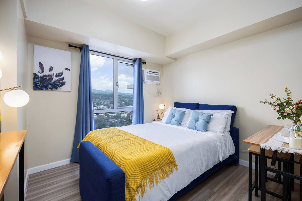 Un dormitorio con cama, escritorio, silla, ventana con cortinas, aire acondicionado y un cuadro en la pared.