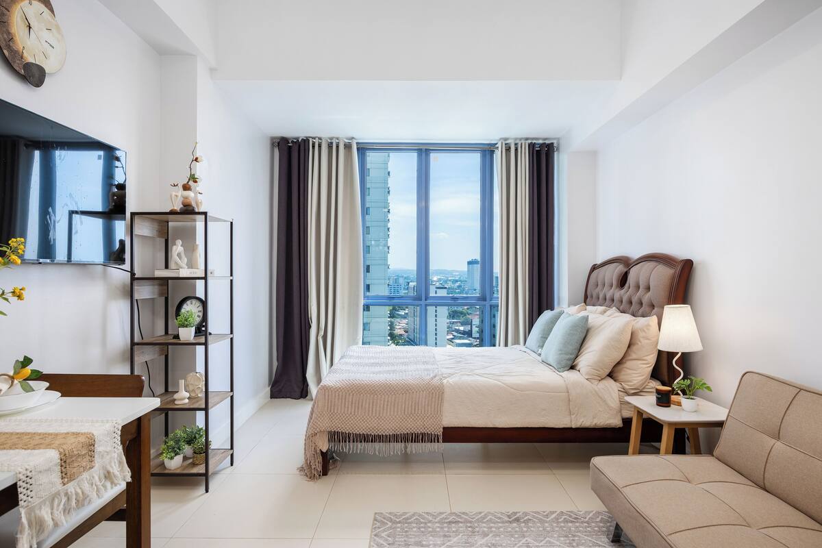 Un dormitorio moderno con un ventanal grande, una cama con cabecera, una mesita de noche con lámpara, un sofá, un estante de madera con objetos decorativos y una pequeña mesa con una planta.