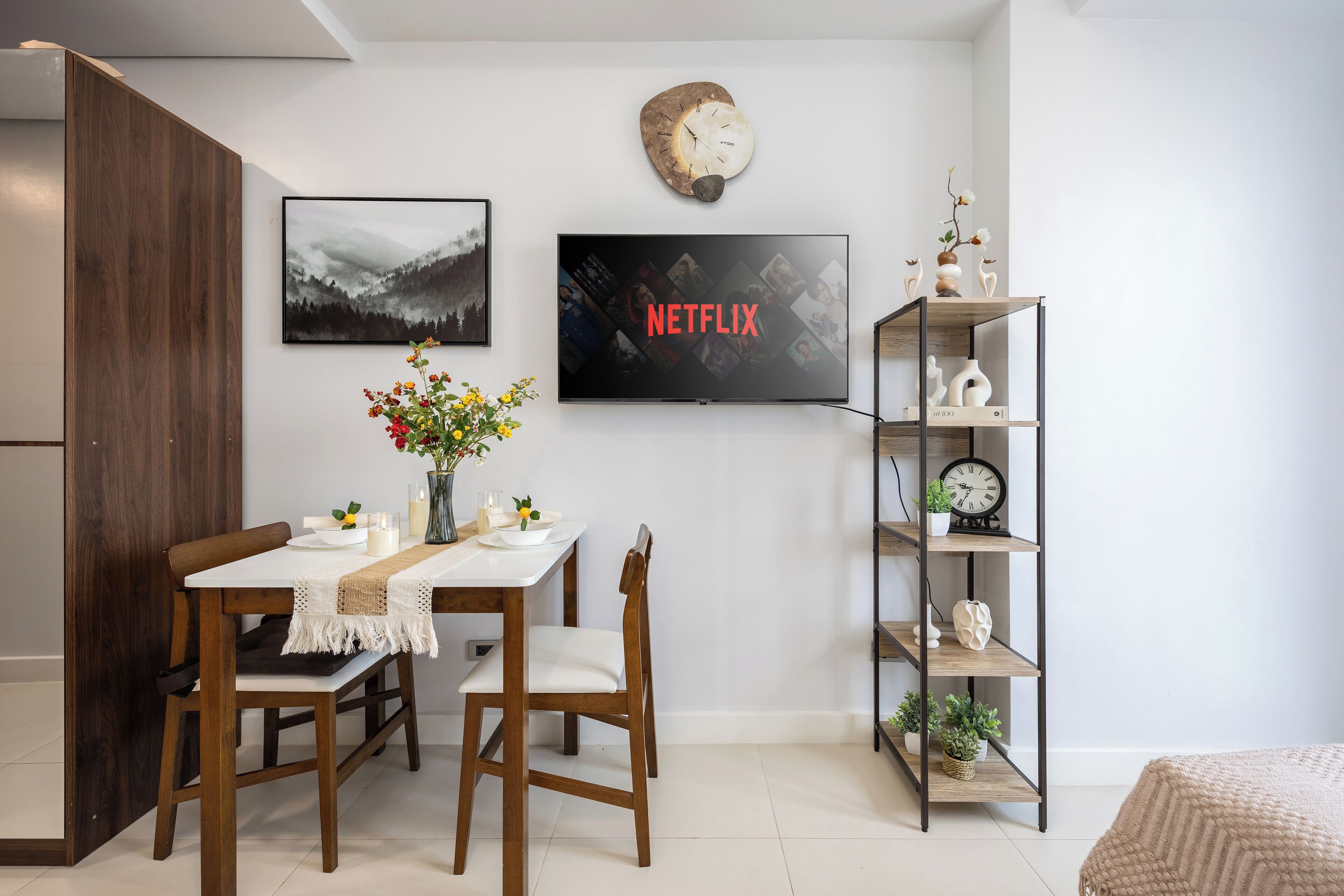 Zona pranzo con tavolo apparecchiato per due, TV a parete che mostra il logo di Netflix e libreria con oggetti decorativi.
