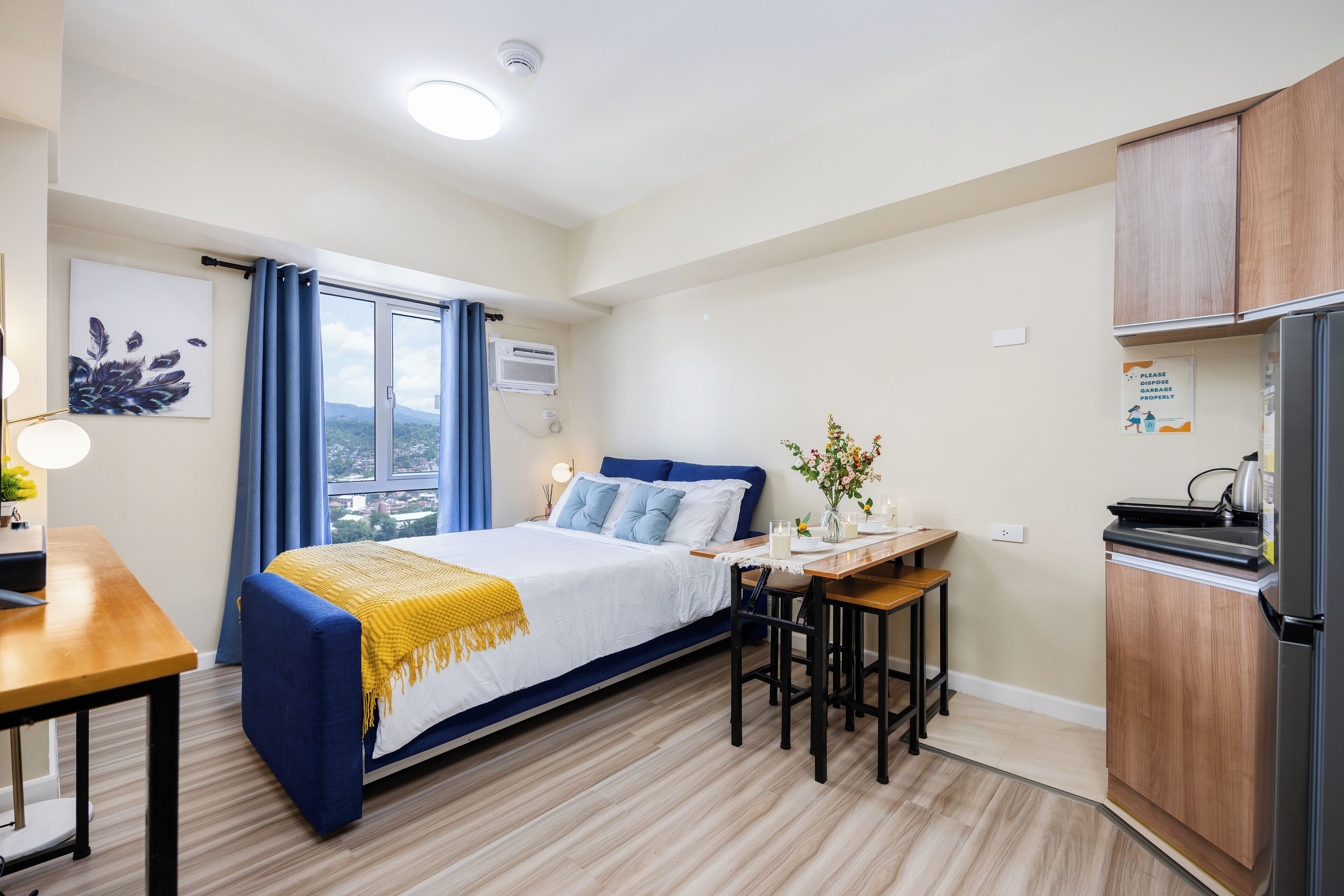 A bedroom with a bed, bedside table, and a small kitchen area with a refrigerator and a sink.