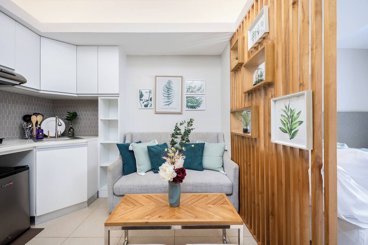 Una cocina moderna con armarios blancos, una mesa de comedor de madera y un sofá gris con cojines turquesa. Hay grabados botánicos enmarcados en la pared y un jarrón con flores sobre la mesa.