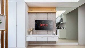 Een moderne woonkamer met een flatscreen televisie aan een muur, een witte tv-kast, en een keuken in de achtergrond.